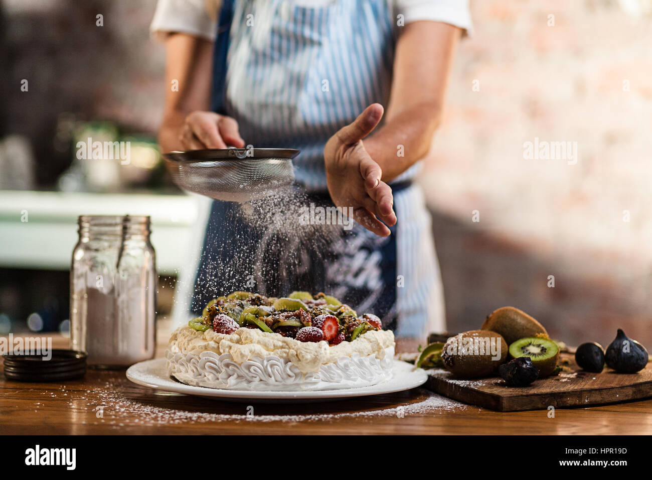 Femme dans une cuisine de style cottage, le tamisage du sucre à glacer sur une pavlova dessert avec un effet de flou sur le sucre glace. Banque D'Images