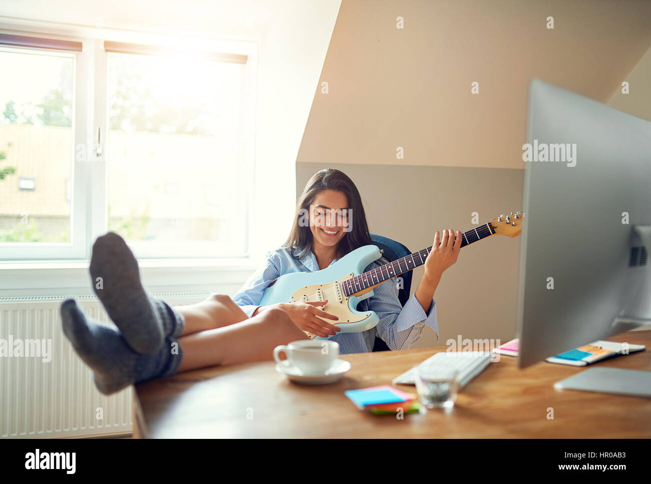 Jolie jeune femme seule à rire avec les pieds sur la table tout en jouant de la guitare électrique en face de moniteur informatique Banque D'Images