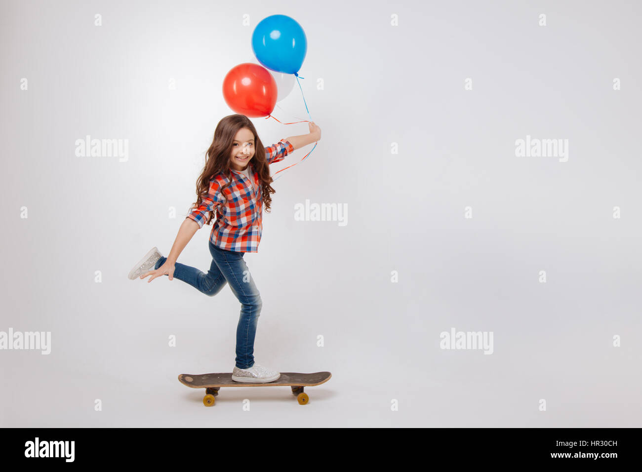 Partager atmosphère positive. Artistique qualifiée Active girl smiling and holding balloons debout contre fond blanc et à l'aide de skateboard Banque D'Images