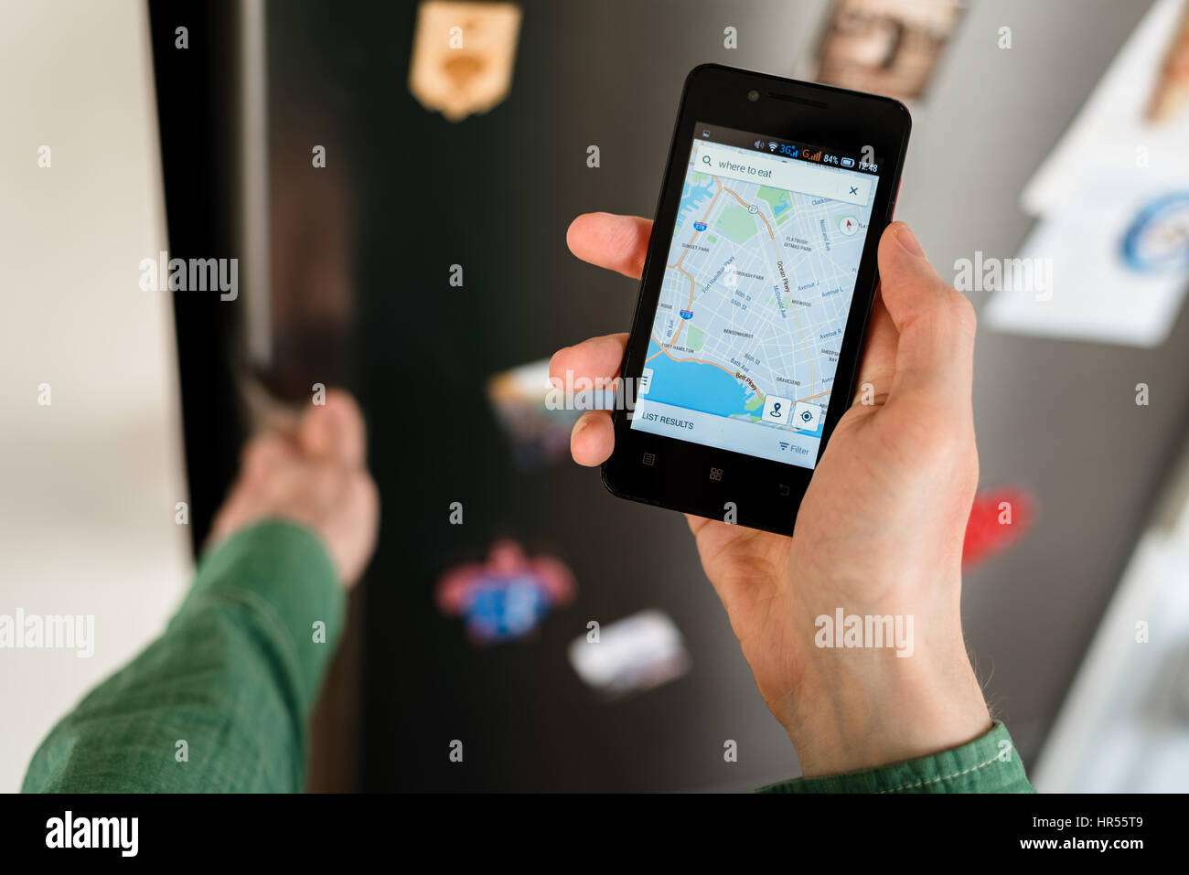 Close-up of personne mains holding smartphone montrant plan de ville à l'écran et d'une recherche en ligne pour les endroits où manger sur cette carte. Réfrigérateur avec aimants o Banque D'Images