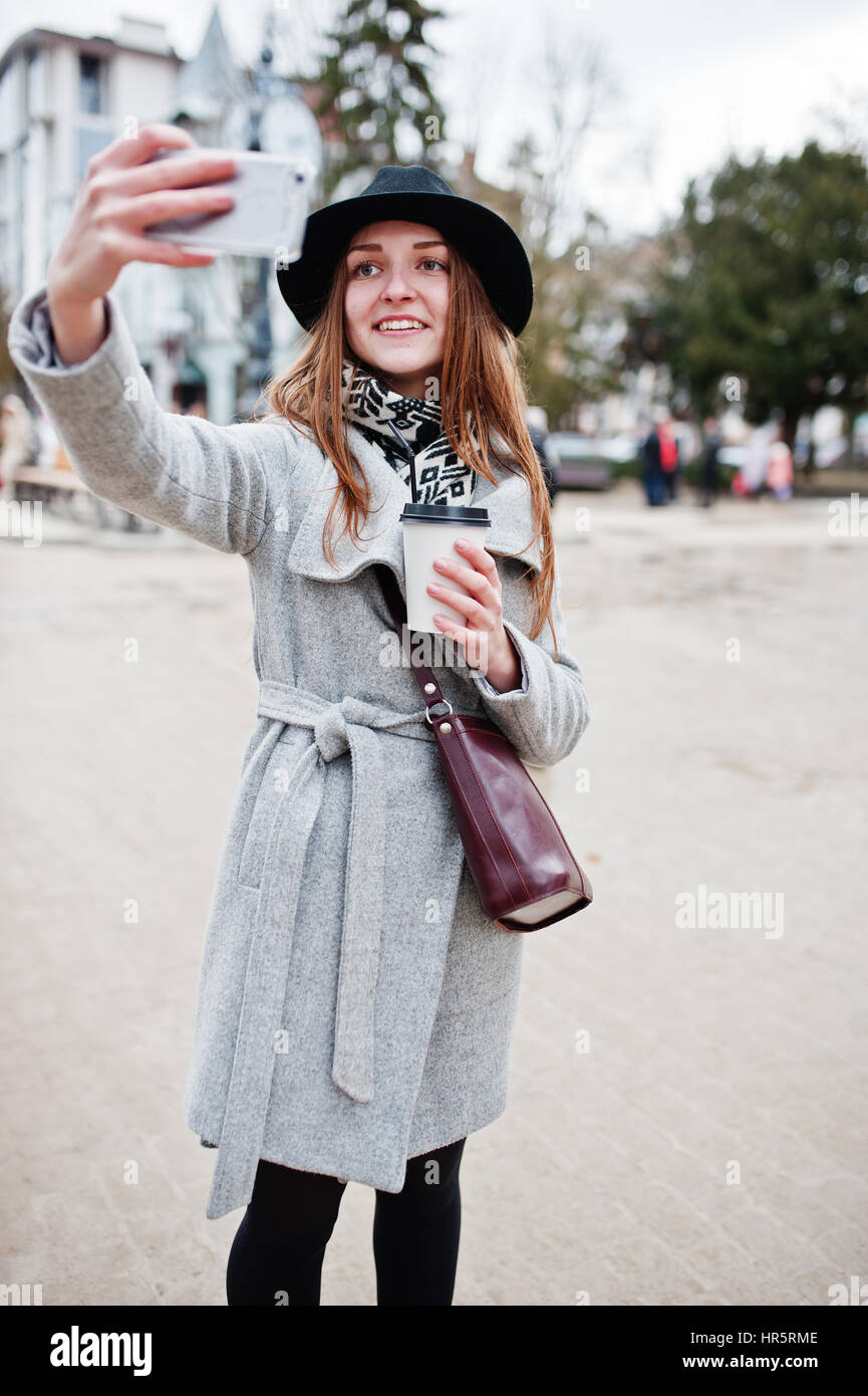 Modèle jeune fille dans un manteau gris et noir chapeau avec sac à main en cuir sur les épaules restent avec tasse de café en plastique et faire en selfies rue de ville. Banque D'Images