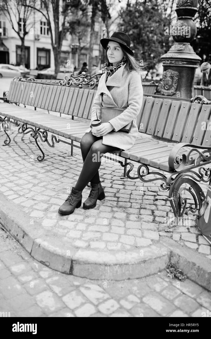 Modèle jeune fille dans un manteau gris et noir chapeau avec sac à main en cuir sur les épaules assis sur un banc avec du plastique tasse de café à la rue de ville. Un noir Banque D'Images