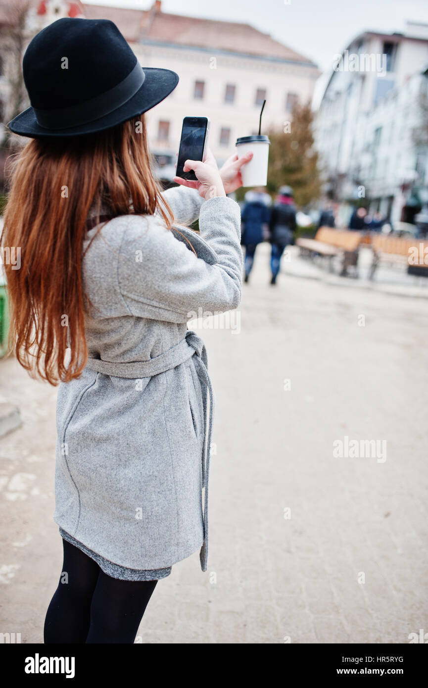 Modèle jeune fille dans un manteau gris et noir chapeau avec sac à main en cuir sur les épaules restent avec tasse de café en plastique et faire en selfies rue de ville. Banque D'Images