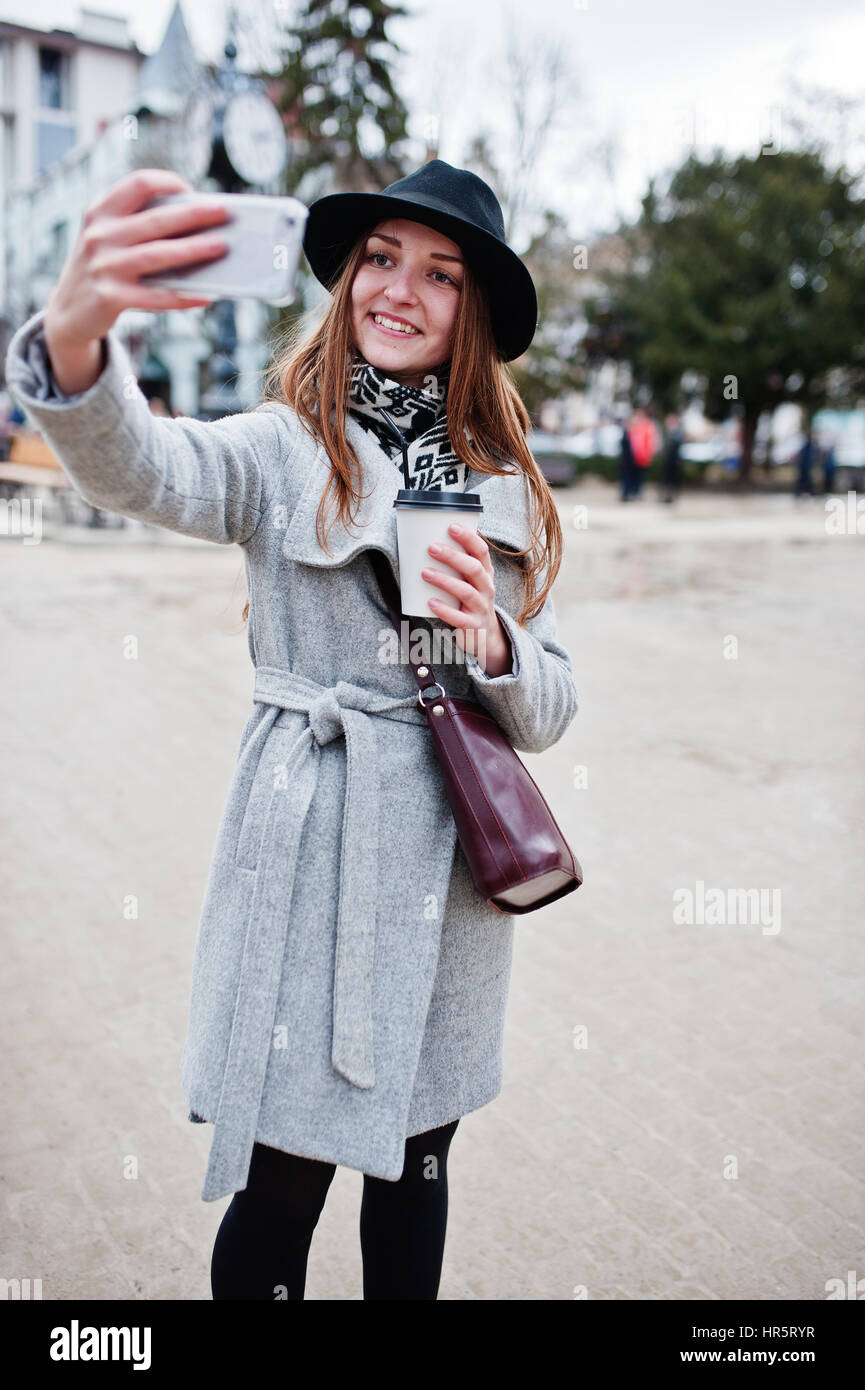 Modèle jeune fille dans un manteau gris et noir chapeau avec sac à main en  cuir sur les épaules restent avec tasse de café en plastique et faire en  selfies rue de