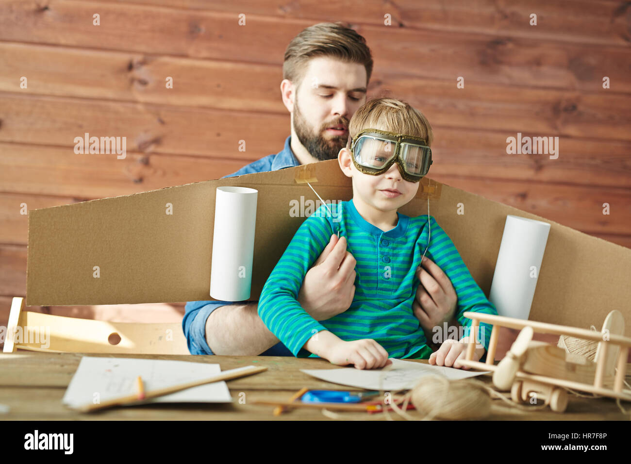 Portrait de jeune père barbu aider son petit-fils en lunettes aviator mettant les cartons d'ailes d'avion Banque D'Images
