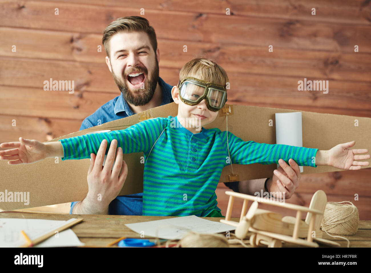 Portrait drôle de père barbu et petit fils de lunettes aviator et carton avec des ailes d'avion derrière son dos jouer ensemble Banque D'Images