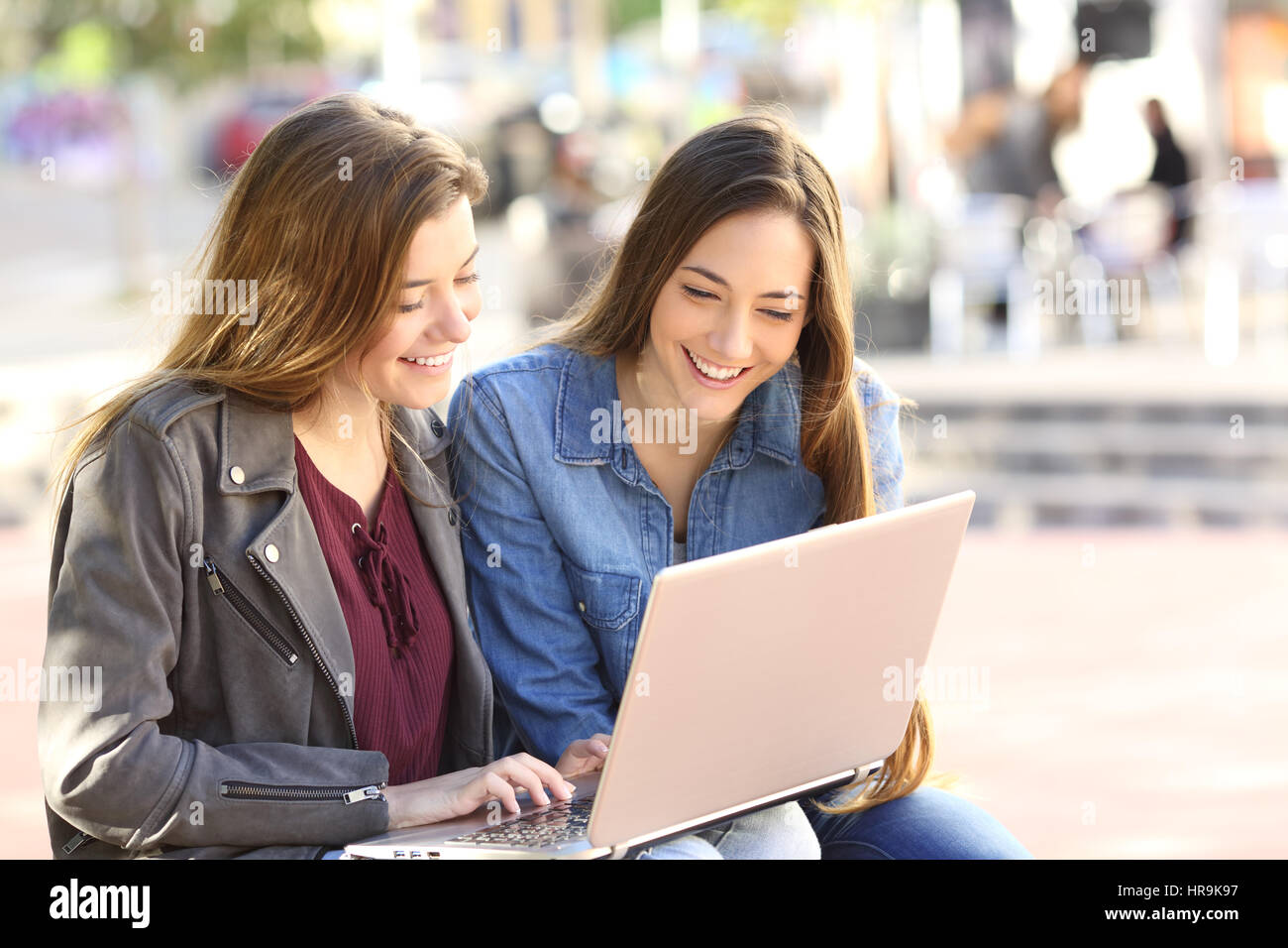 Deux amis de regarder du contenu multimédia en ligne avec un ordinateur portable assis sur un banc dans la rue avec un milieu urbain Banque D'Images