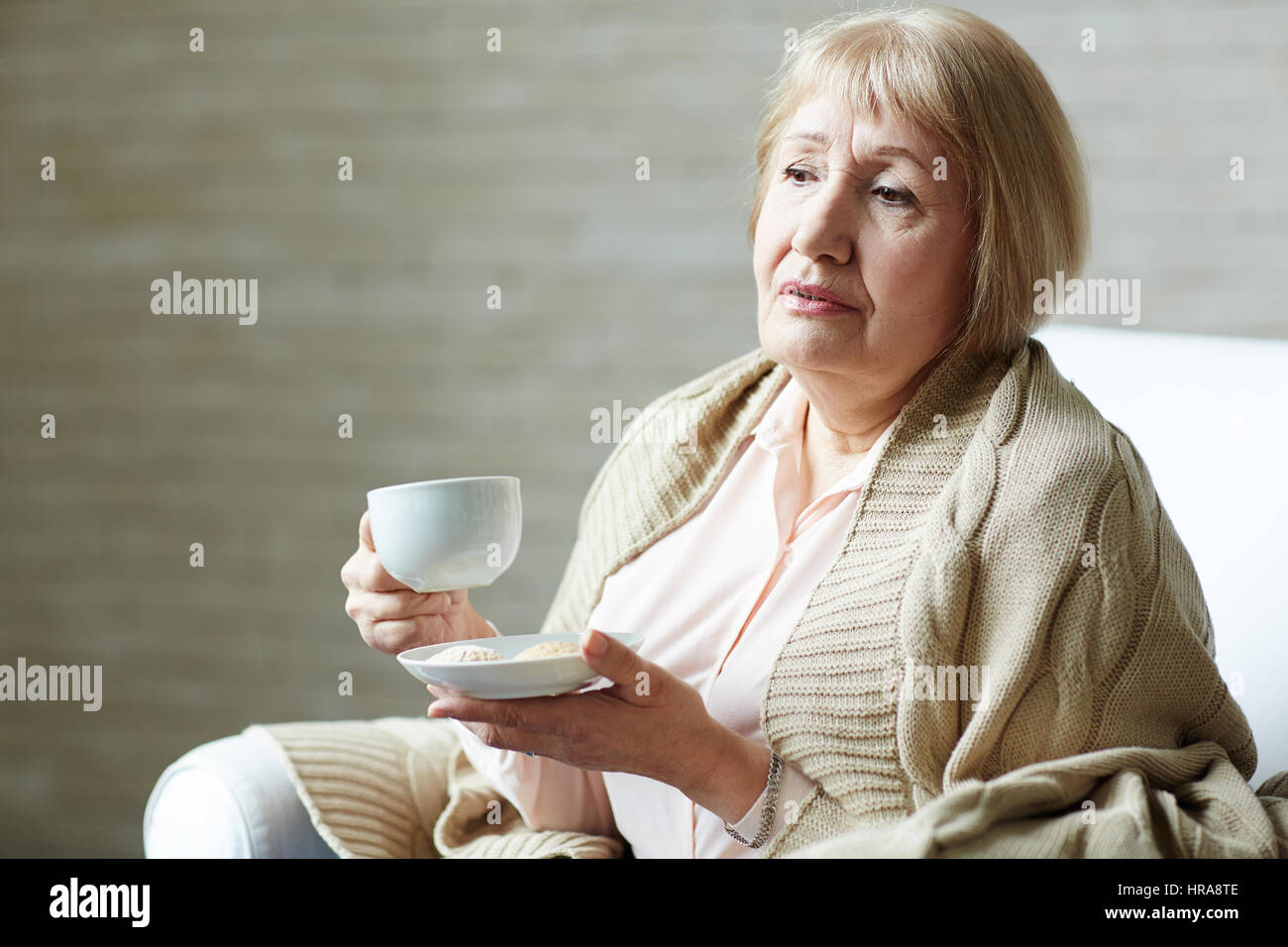 Senior femme mélancolique avec plaid sur les épaules holding coffee mug dans une main et une soucoupe avec des cookies dans d'autres tout en vous relaxant dans la salle de séjour Banque D'Images