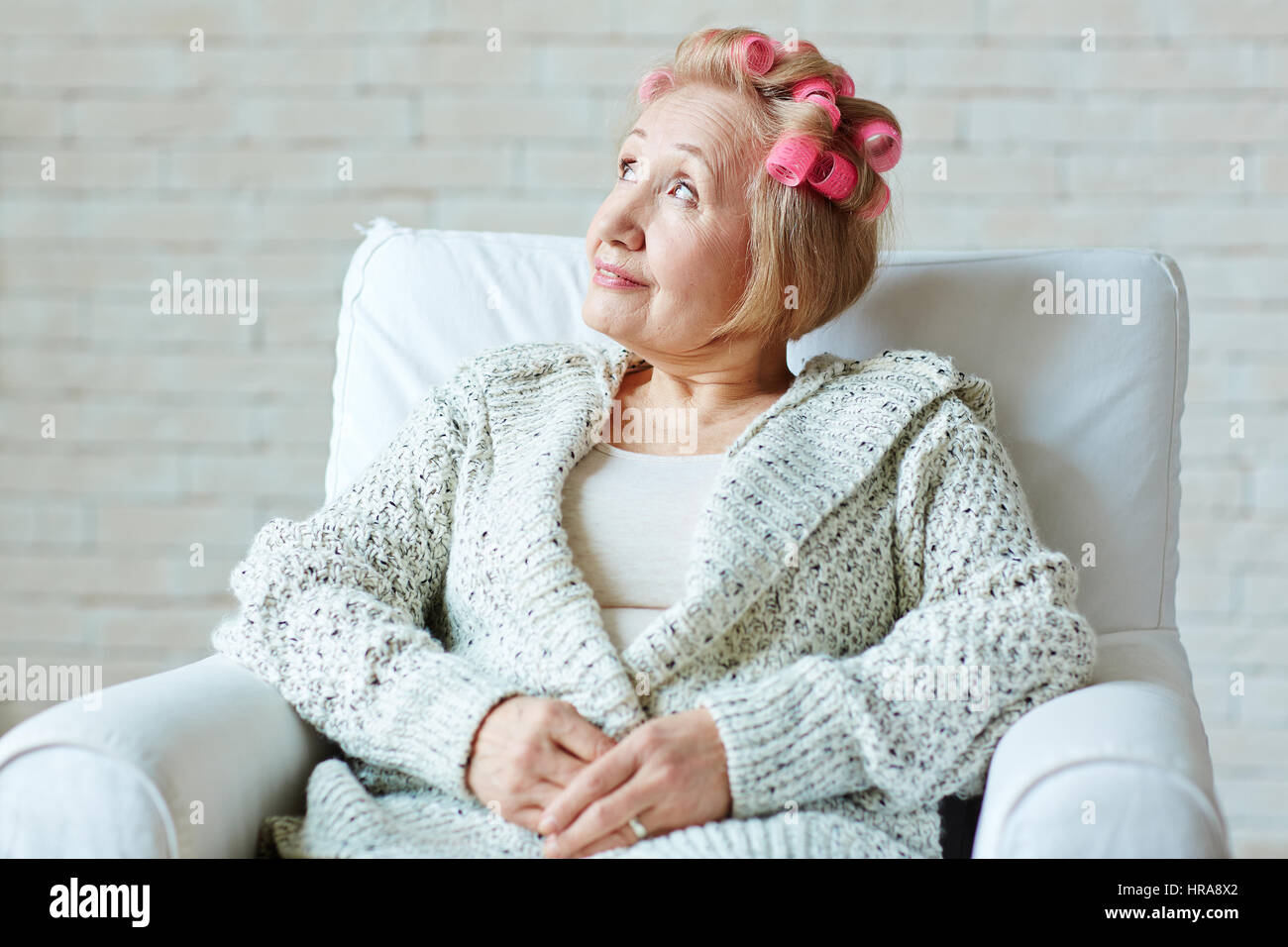 Portrait of senior woman de rêve avec hair rollers et dans des étoffes cardigan blanc assis sur un fauteuil et regardant vers le haut avec le sourire Banque D'Images