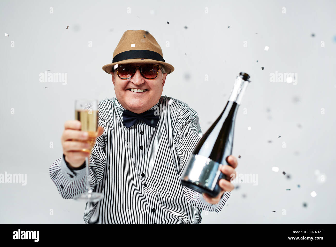 Jusqu'à la taille portrait de vieil homme excité en chapeau melon et des lunettes acclamer avec bouteille de champagne et confettis Banque D'Images