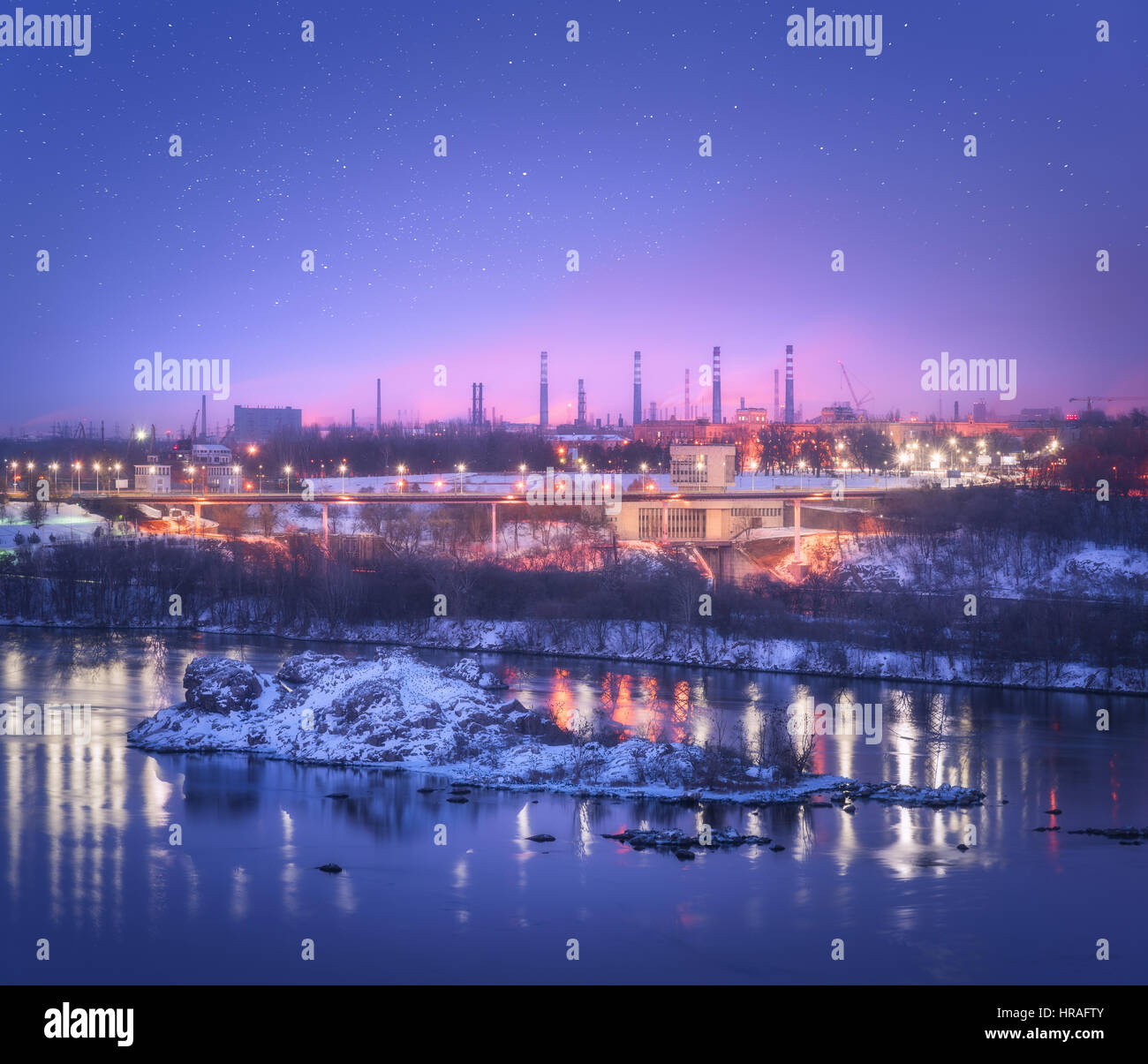 Nuit paysage urbain avec purple sky coloré avec des étoiles, rochers, rivière, arbres, bâtiments, illumination de ville et de l'acier usine avec cheminée en hiver Banque D'Images
