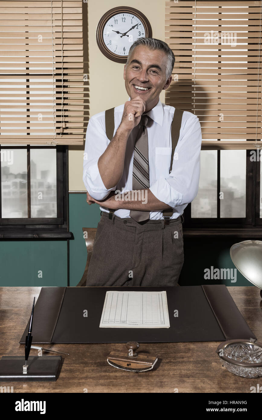 Attractive businessman with hand on chin smiling at camera, bureau vintage. Banque D'Images Attractive businessman with hand on chin smiling at camera, bureau vintage. Banque D'Images