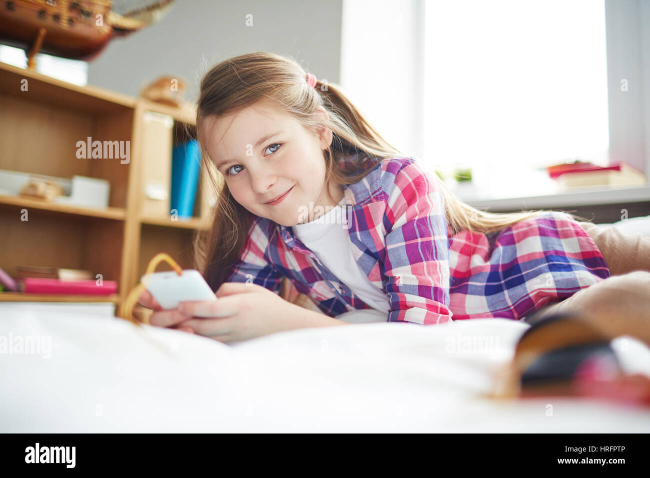 Close-up portrait of pretty little Girl with ponytails lying on bed, holding smartphone and smiling at camera gaiement Banque D'Images