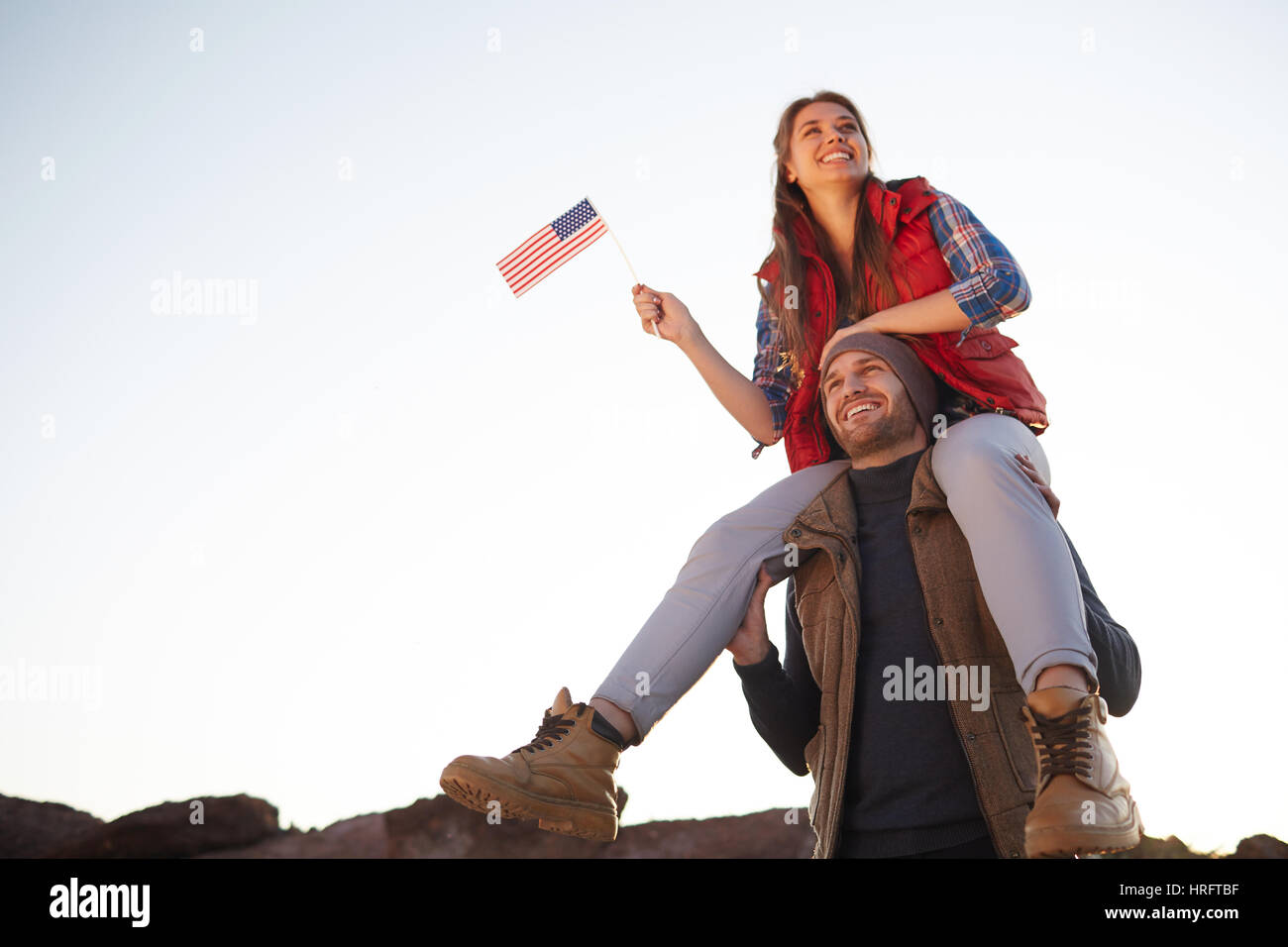 Low angle portrait of young tourist couple célébrant la liberté américaine : beautiful girl sitting on her boyfriends épaules en souriant gaiement et wav Banque D'Images