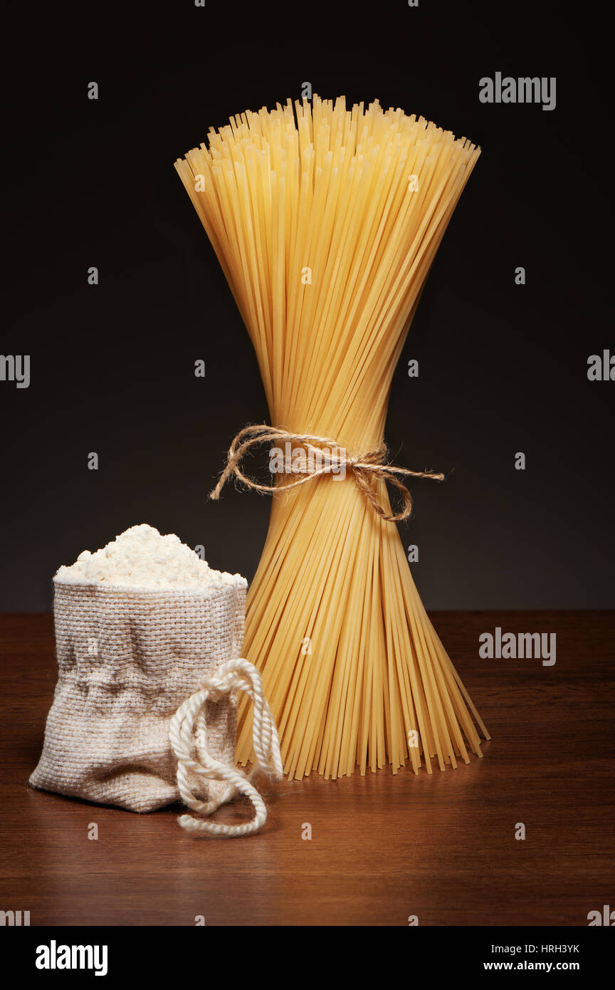 Les pâtes spaghetti sec attaché avec de la corde et de la toile de jute sac de farine sur la table en bois sombre sur fond gris Banque D'Images