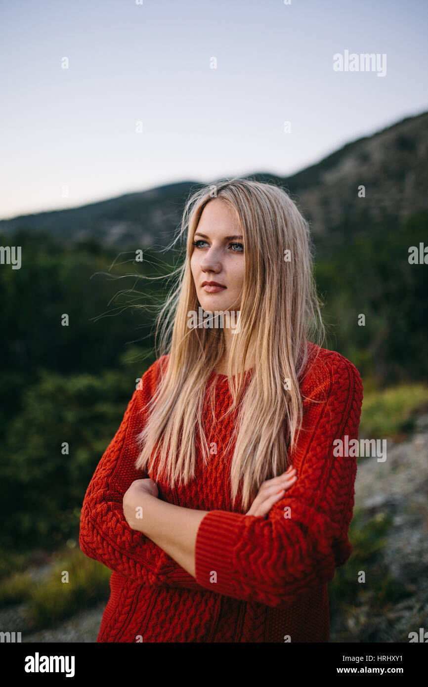 Portrait of a young blonde woman in red pullover avec bras croisés en plein air, sur l'arrière-plan de montagnes et des forêts Banque D'Images