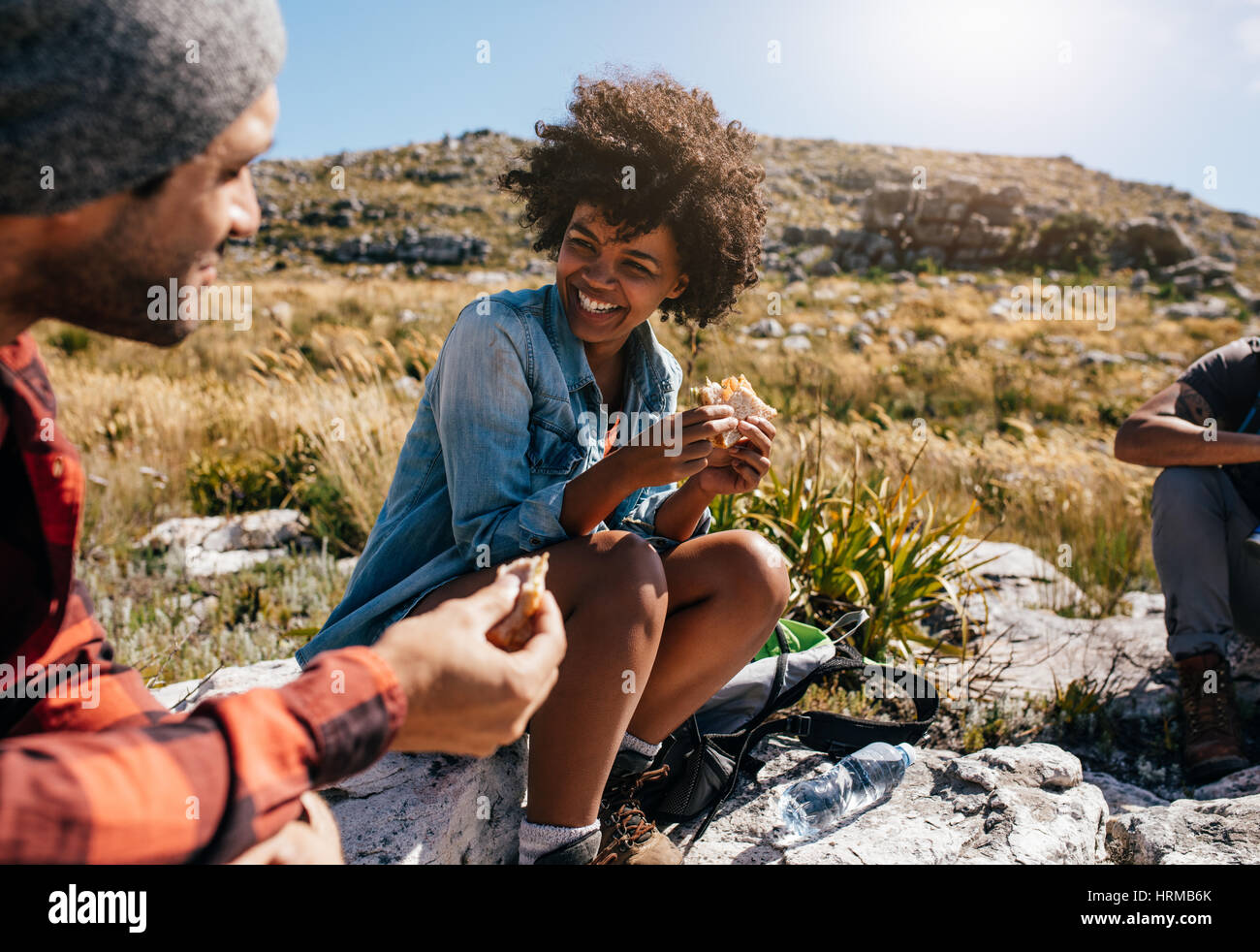 Happy young African woman eating sandwich et souriant tout en vous relaxant avec des amis au cours de la randonnée. Groupe de randonneurs en pause pendant la randonnée de pays. Banque D'Images