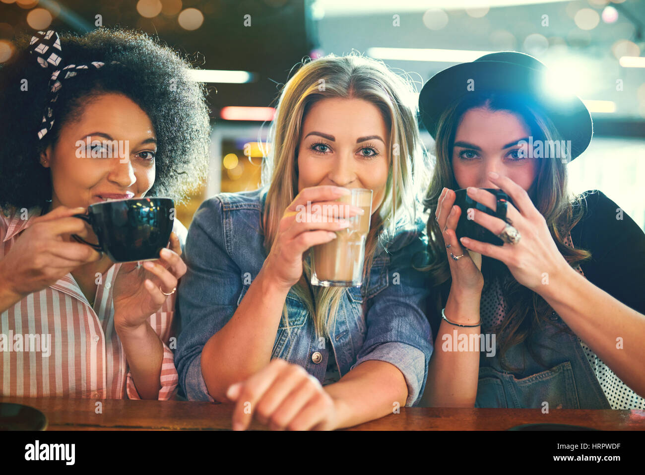 Trois jeunes femmes/portrait close-up au cafe table sirotant du café à partir de leurs tasses, looking at camera and smiling Banque D'Images