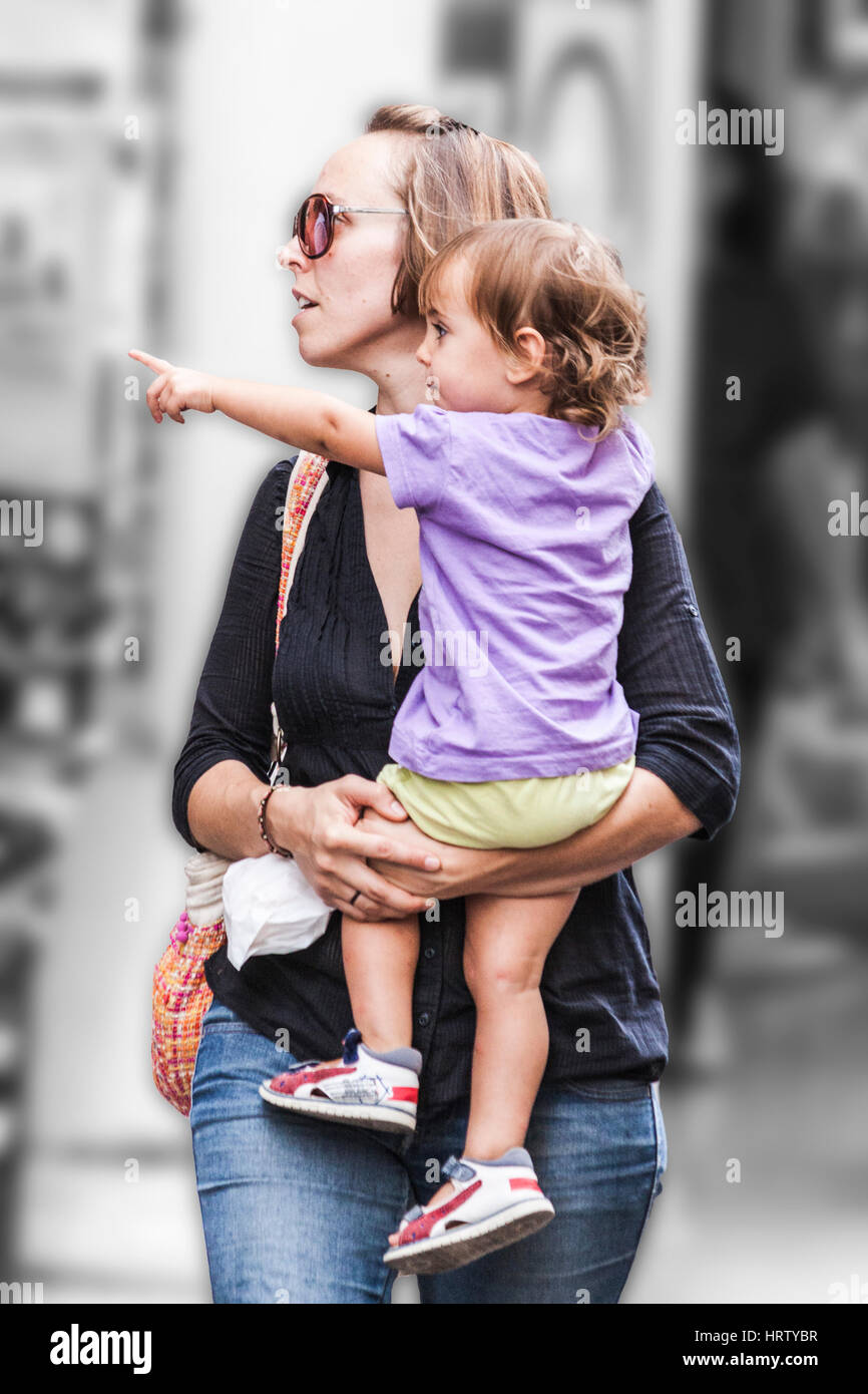 Maman marche sur la rue et porte le jeune enfant qui indique quelque chose qu'elle a remarqué Banque D'Images