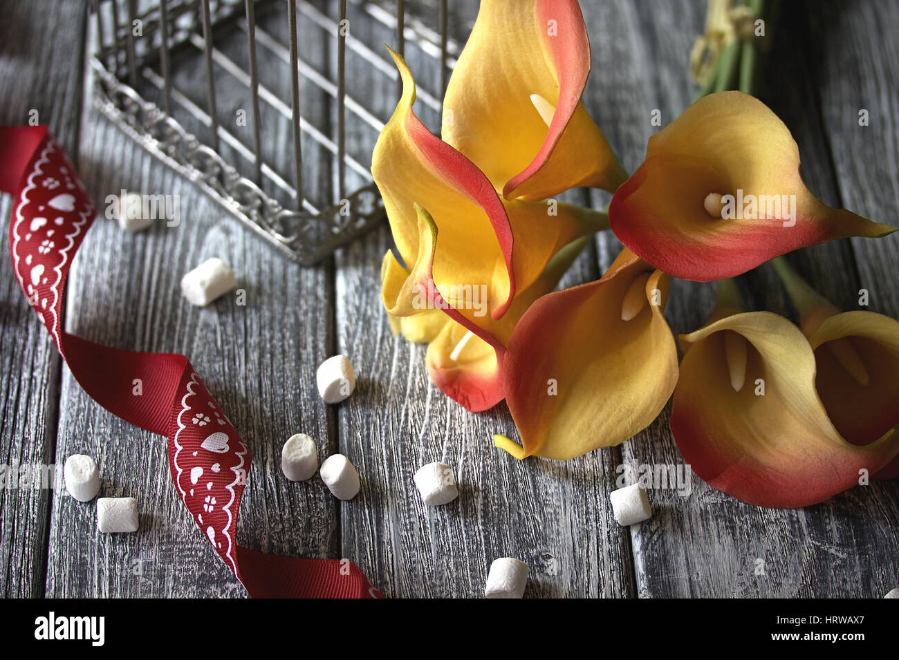 Des lis calla jaune orange avec ruban rouge et blanc des guimauves sur fond gris en bois Banque D'Images