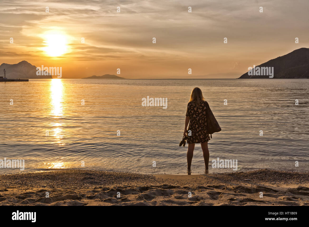 Belle jeune femme, pieds nus et avec de longs cheveux blonds regardant le coucher du soleil doré sur une île grecque, Amorgos Banque D'Images