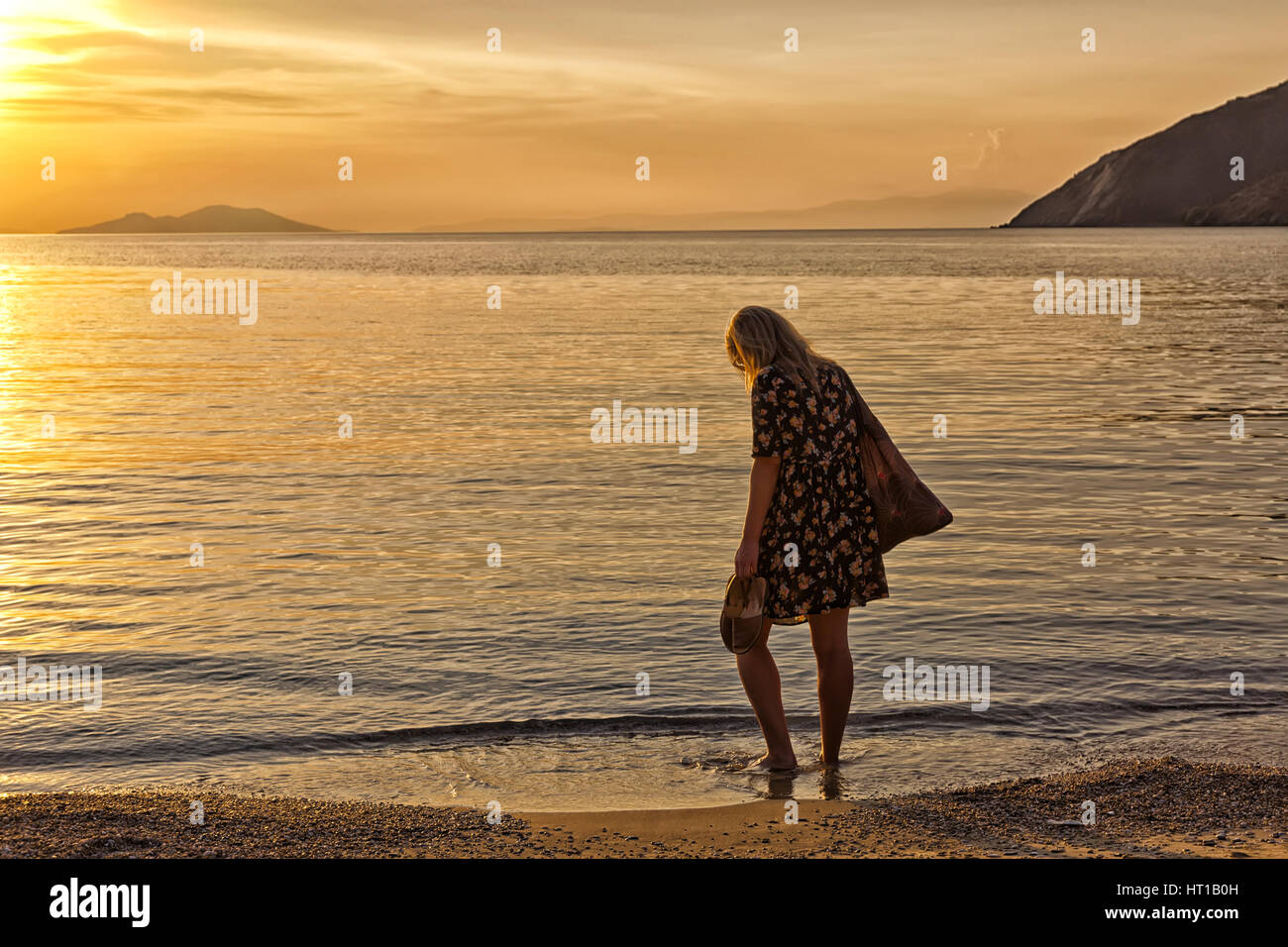 Belle jeune femme, pieds nus et avec de longs cheveux blonds regardant le coucher du soleil doré sur une île grecque, Amorgos Banque D'Images