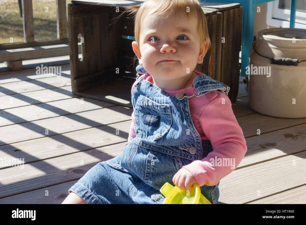 Un an smiling Caucasian baby girl en salopette assis sur une terrasse extérieure avec un verre tasse. New York, USA. Banque D'Images