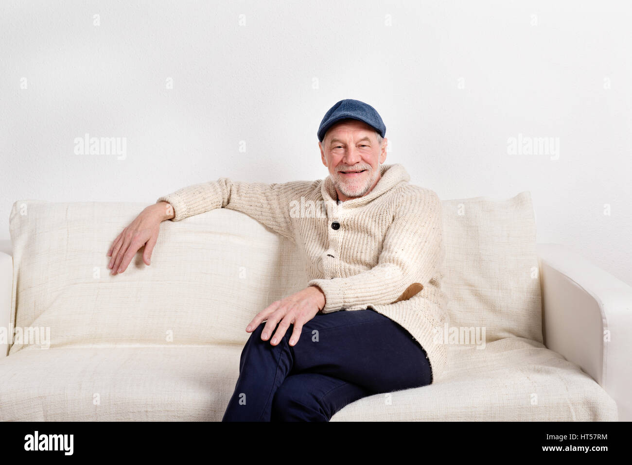 Handsome man pull beige en souriant, assis sur un canapé, les jambes croisées. Studio shot contre le mur blanc. Banque D'Images