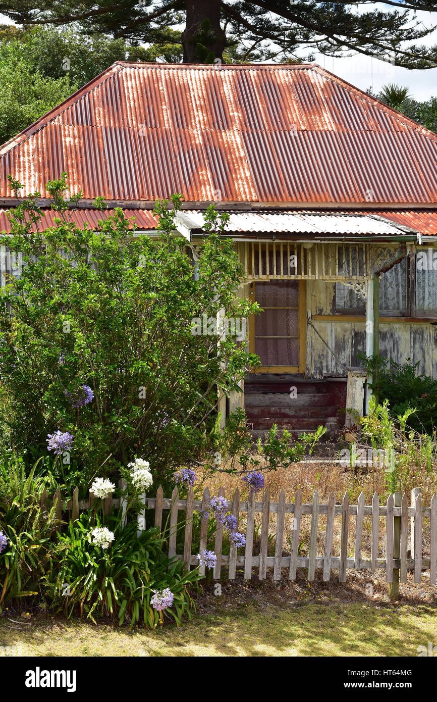 Maison en bois patiné très en forme de sable avec toit de tôle ondulée rouillée dans la verdure derrière la petite barrière en bois. Banque D'Images