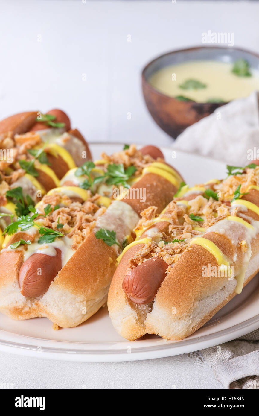 Hot dog avec de la saucisse, oignon frit, des feuilles de coriandre, de moutarde et de la sauce au fromage blanc, servi sur une plaque en céramique avec plus de textile texture béton blanc Banque D'Images