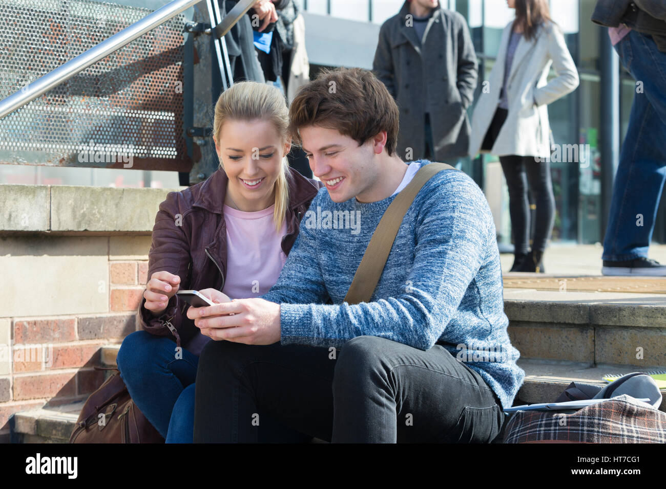 Un jeune couple sympathique sourire alors qu'ils s'assoient sur des pas dans la ville à la recherche d'un téléphone mobile. Il y a un groupe d'autres étudiants de se concentrer dans le bac Banque D'Images