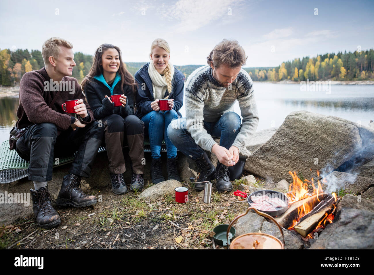 Jeunes amis le café tout en regardant l'homme à la cuisson des aliments au camping au bord du lac Banque D'Images