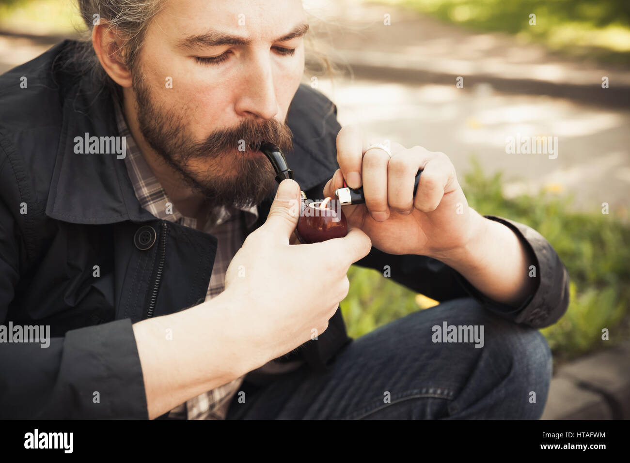 Jeune homme barbu fumant une pipe en parc d'été, close-up portrait with selective focus Banque D'Images