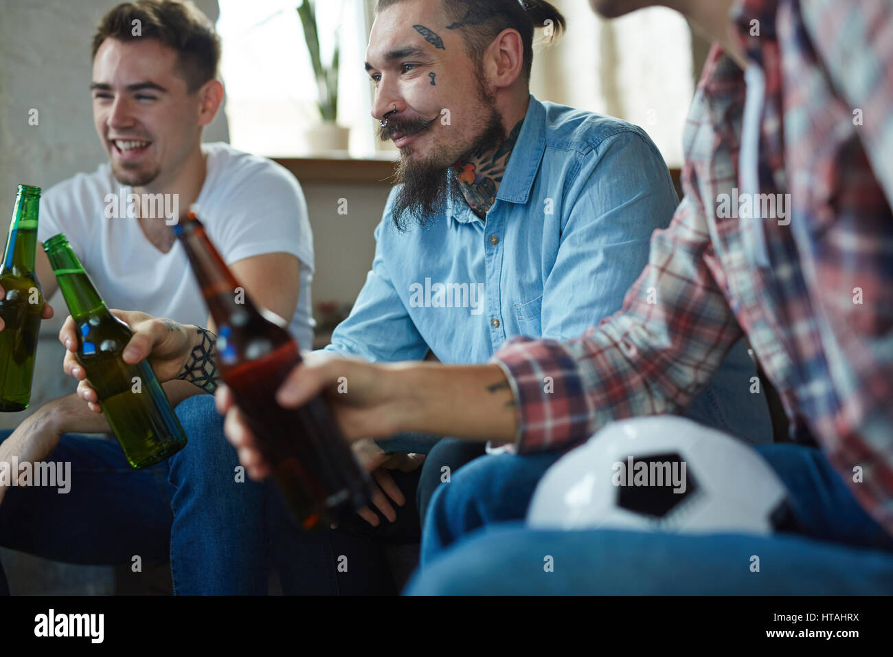 Guy avec barbe et tatouages holding bouteille de bière en regardant la télévision football Banque D'Images