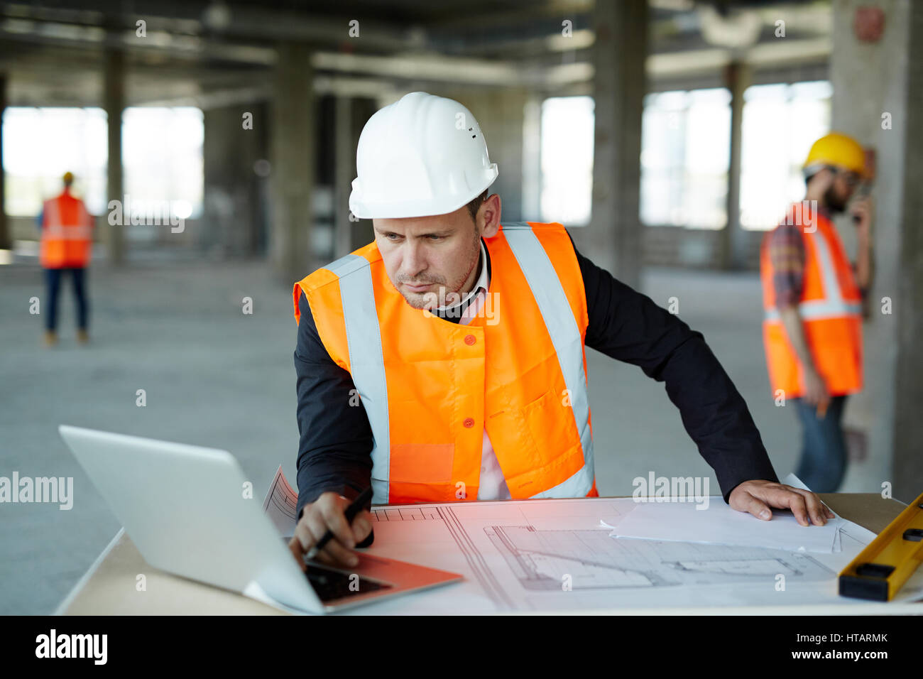 Portrait de superviseur développement le port de casque et gilet costume formel sur les plans d'épreuvage à l'aide d'un ordinateur portable à l'intérieur unfinished bu Banque D'Images