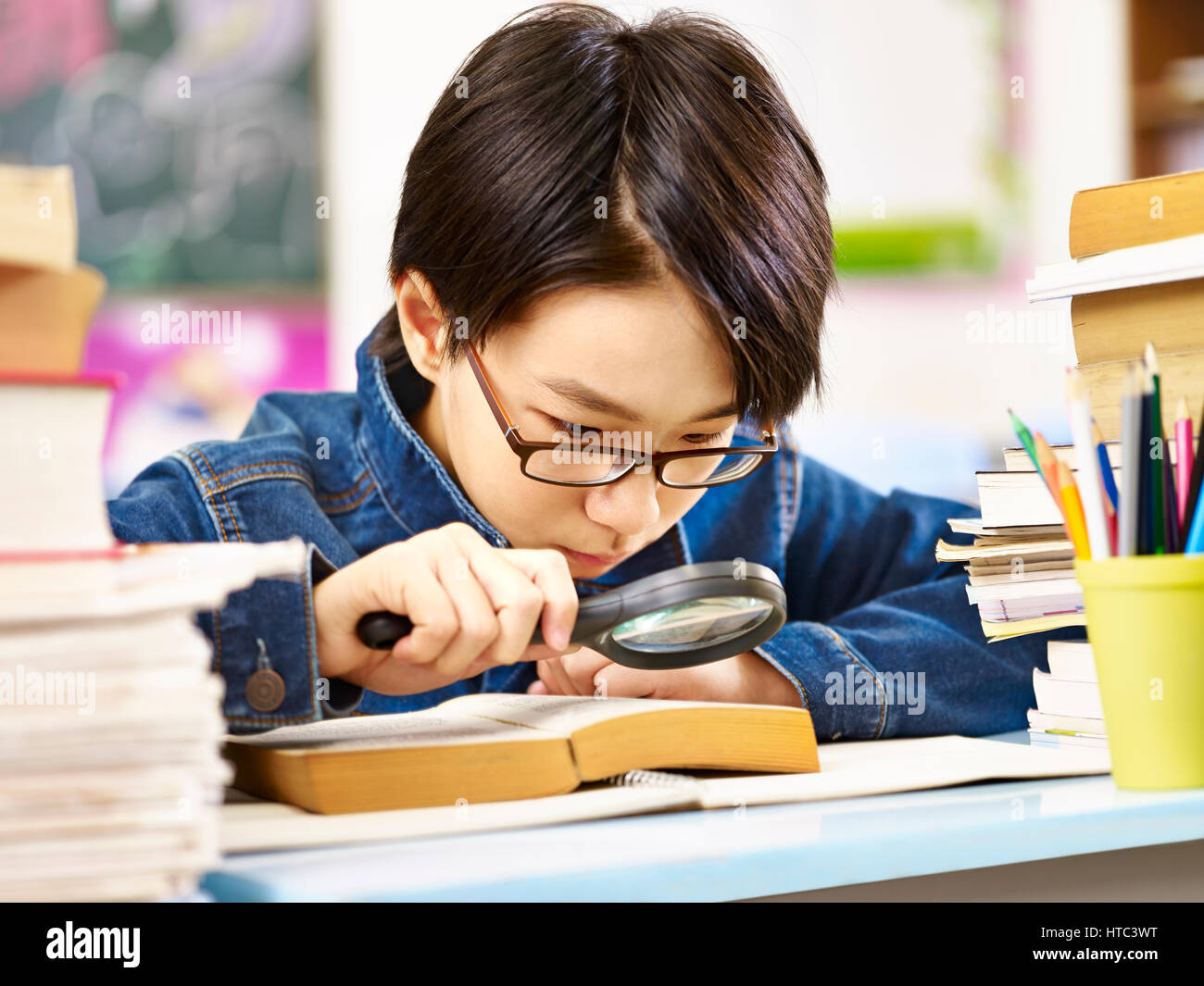 Élève d'Asie avec des lunettes à l'aide d'une loupe pour agrandir les mots dans un livre épais. Banque D'Images