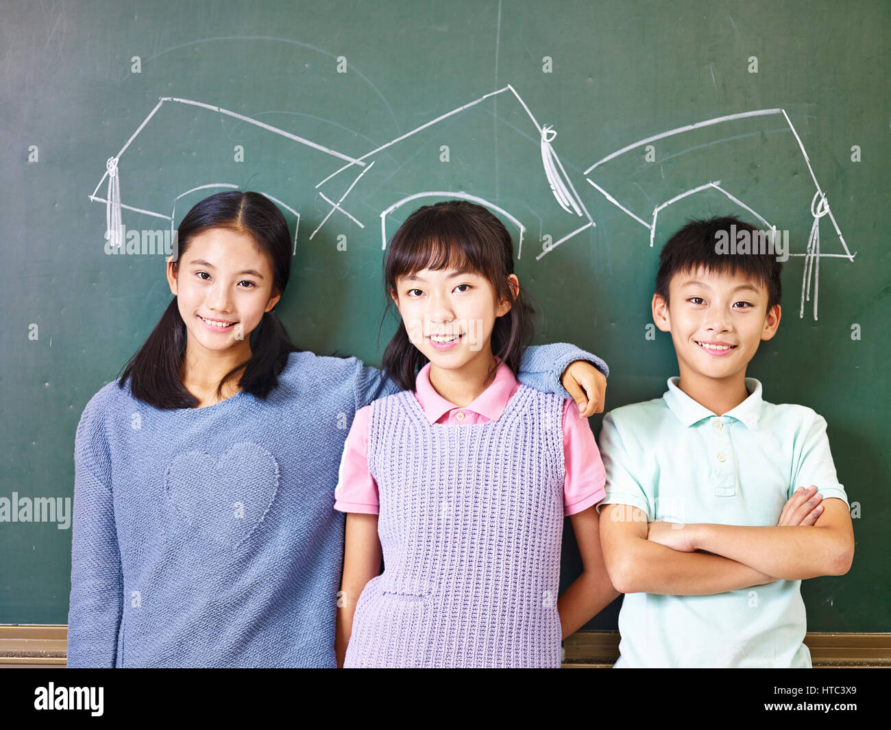 Trois enfants de l'école primaire asiatique debout sous la craie-tirées des chapeaux de doctorat in front of blackboard. Banque D'Images