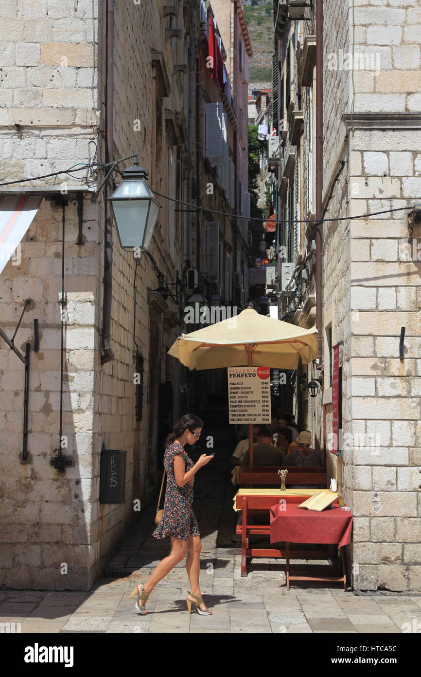 Une jolie jeune femme dans une robe d'été courte les sms sont en marchant au-delà d'un café de la rue dans la ville historique de Dubrovnik, Croatie Banque D'Images Une jolie jeune femme dans une robe d'été courte les sms sont en marchant au-delà d'un café de la rue dans la ville historique de Dubrovnik, Croatie Banque D'Images