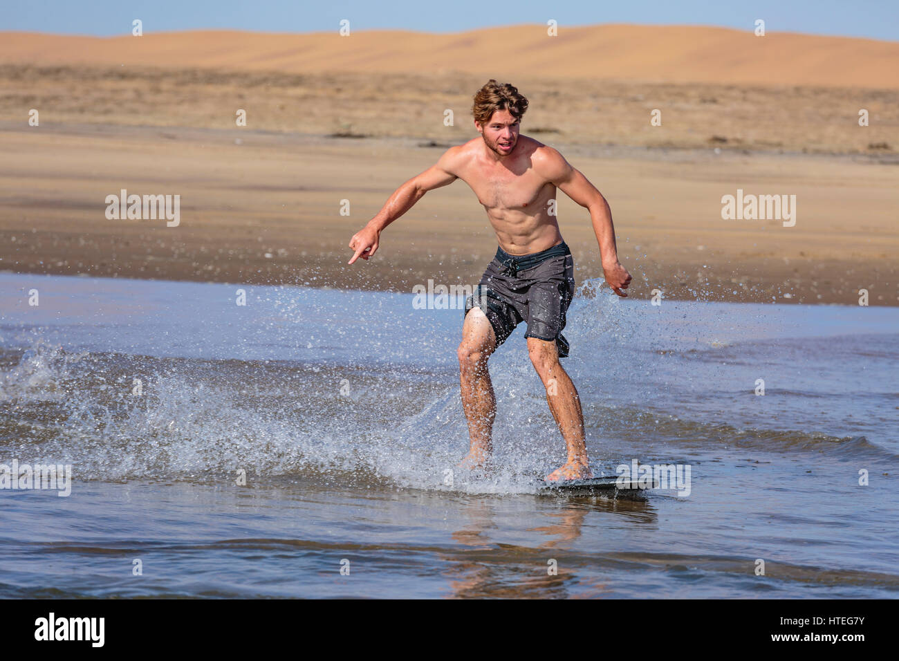 Jeune homme skimboarding dans la mer, à l'arrière des dunes, Swakopmund, Namibie Banque D'Images