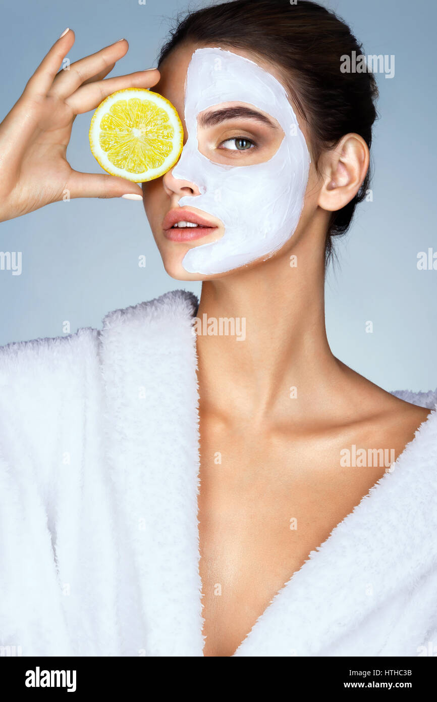 Brunette woman holding d'une tranche de citron en face de son visage. Photo de femme avec masque facial hydratant. Beauté et Soins De La Peau concept Banque D'Images