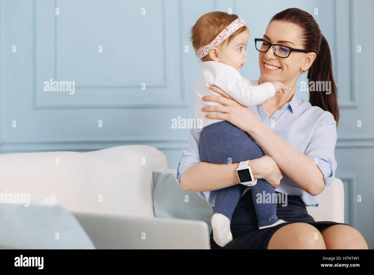Doux et attentionné. Jeune femme énergique accompli assis sur un canapé et de dépenser sa pause avec sa petite fille tout en travaillant de chez vous. Banque D'Images