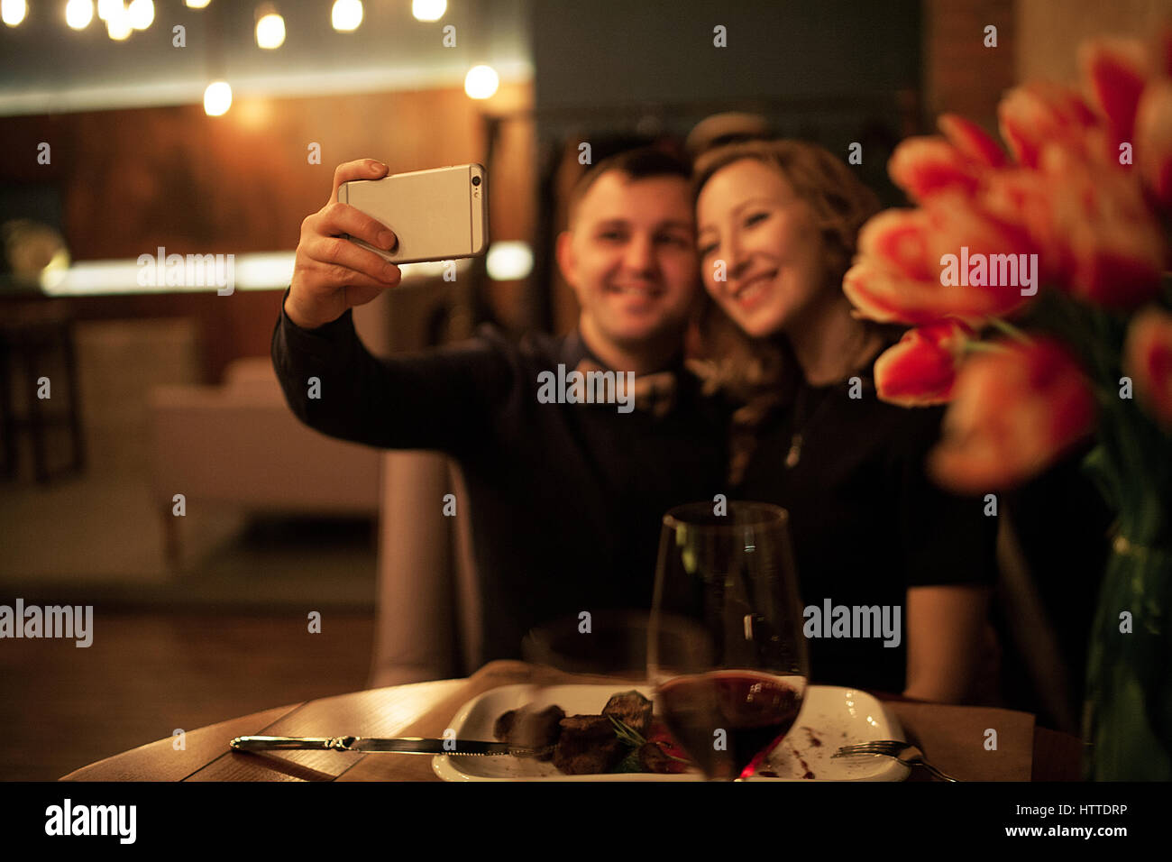 Jeune homme et femme faisant selfy par smartphone dans un restaurant. Avant d'entre eux est le tableau avec des aliments et des verres de vin. Banque D'Images