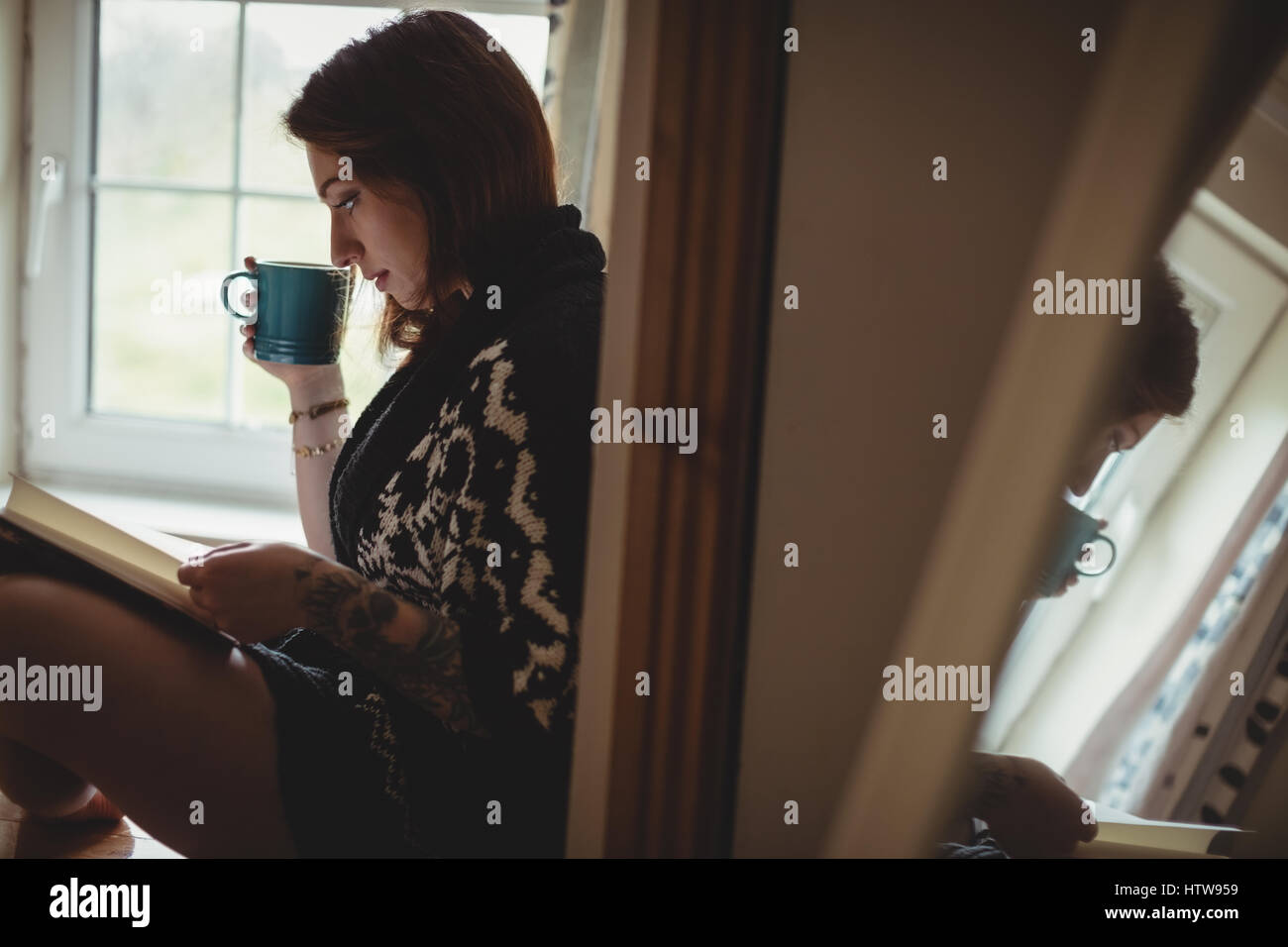 Woman having coffee et la lecture d'un livre tout en restant assis à l'appui de fenêtre Banque D'Images