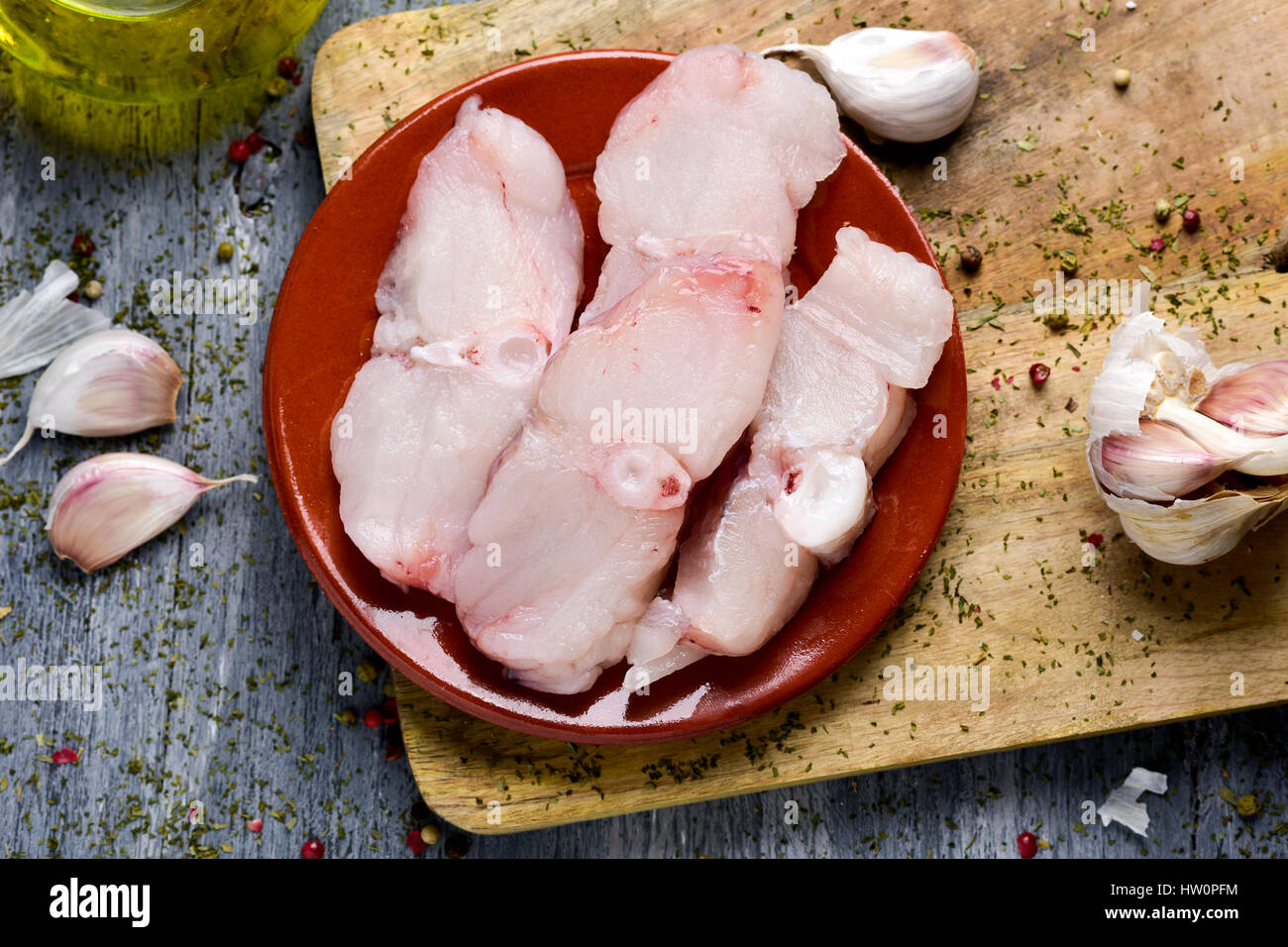 Tourné à angle élevé de certaines tranches de raw monkfish sur un conseil d'achat de bois, quelques ails et une salière de l'huile d'olive sur une table en bois rustique Banque D'Images