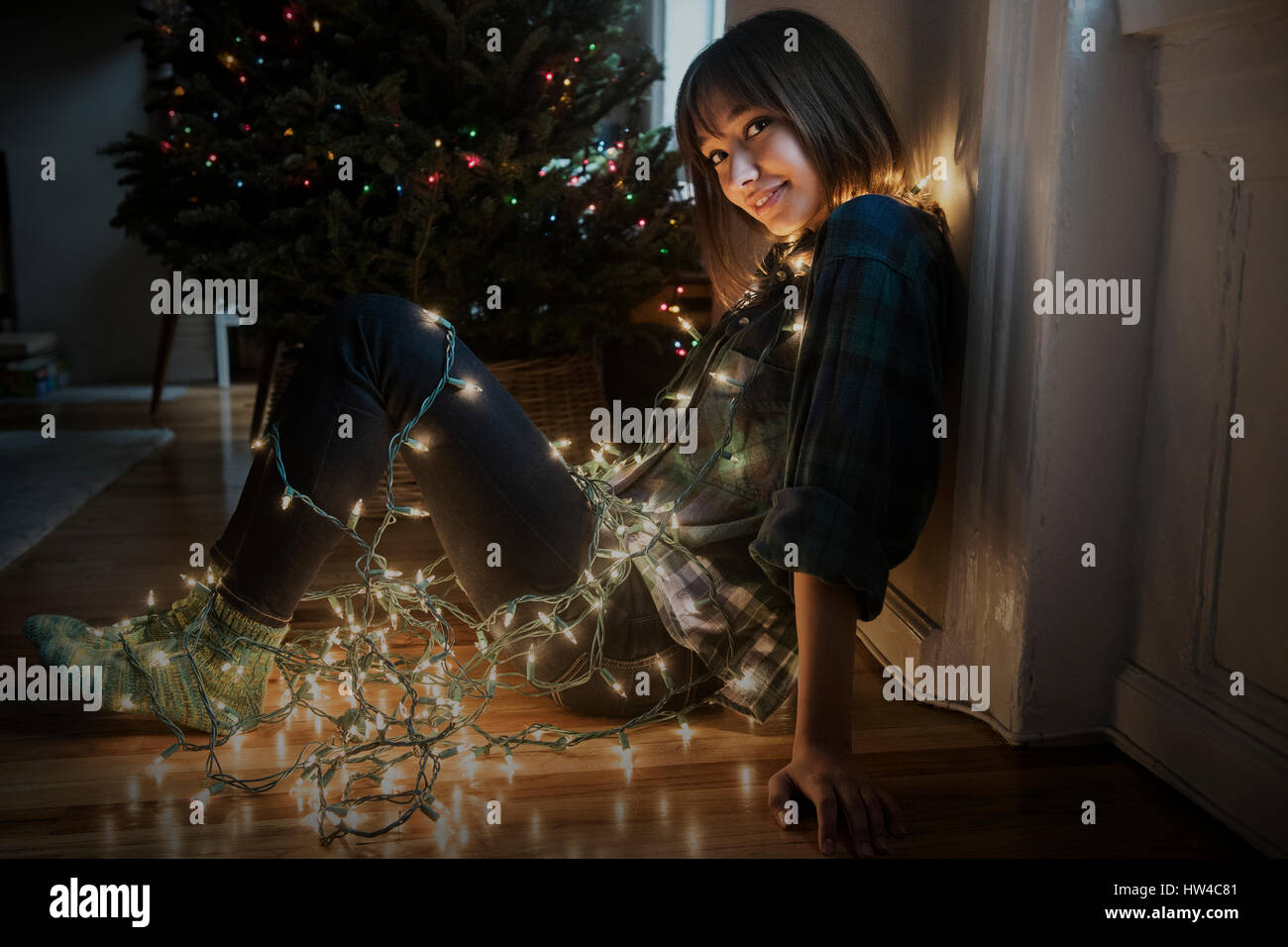 Mixed Race woman sitting on floor enveloppé dans string lights near Christmas Tree Banque D'Images