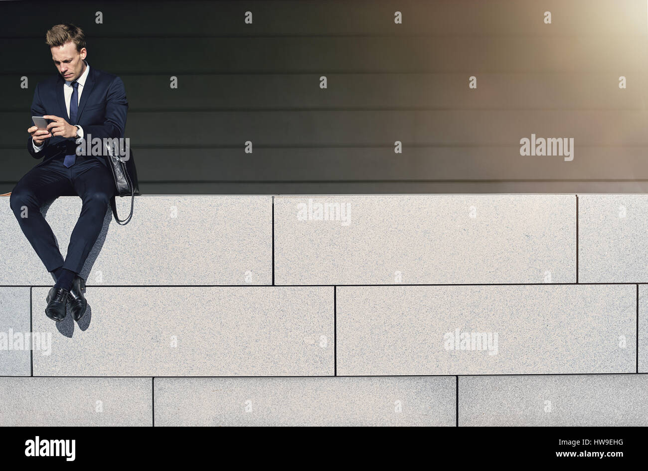 Businessman utilise son smartphone tout en étant assis sur le mur de briques. Tourné en extérieur horizontal Banque D'Images