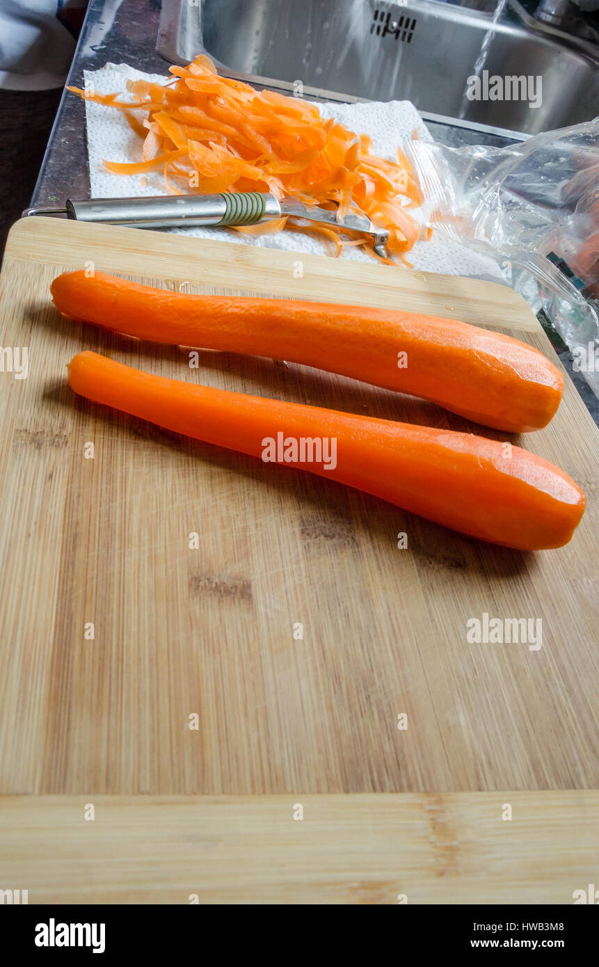 Un couple de carottes épluchées sur une planche à découper en bois dans une cuisine maison. La carotte des peelings et peeler sont sur le plan de travail de cuisine en arrière-plan. Banque D'Images