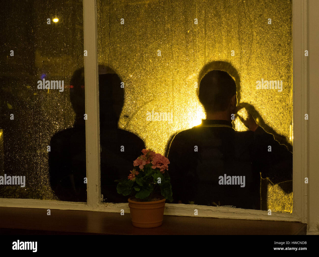 Deux fumeurs anonymes à l'extérieur d'un rétro-éclairé fenêtre pluie avec leurs ombres projetées sur le verre humide par la lumière artificielle d'or Banque D'Images