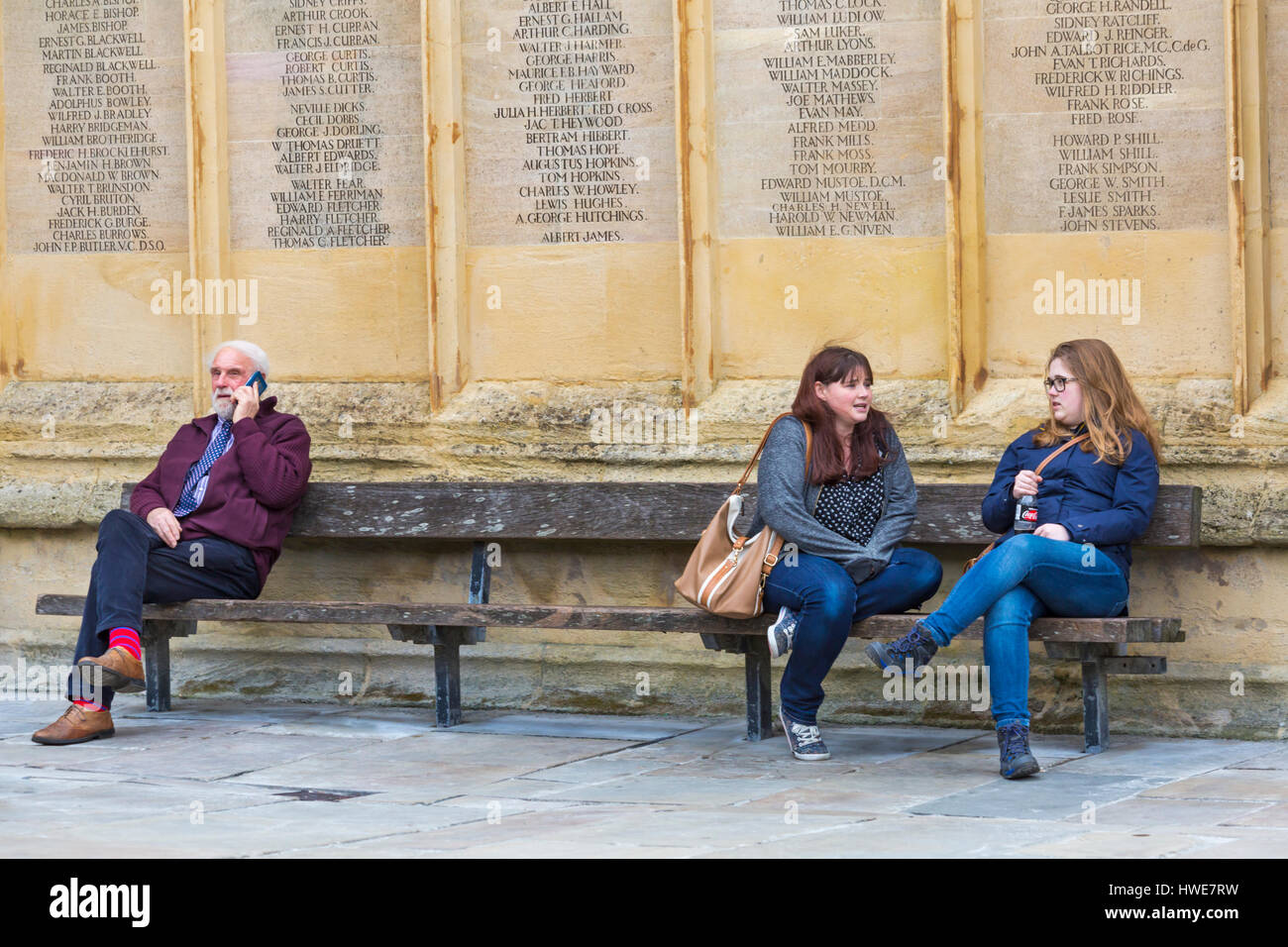 Cirencester - Trois personnes assis sur un banc - deux jeunes femmes bavardant tandis que man talks on mobile phone at Cirencester, Gloucestershire en Mars Banque D'Images