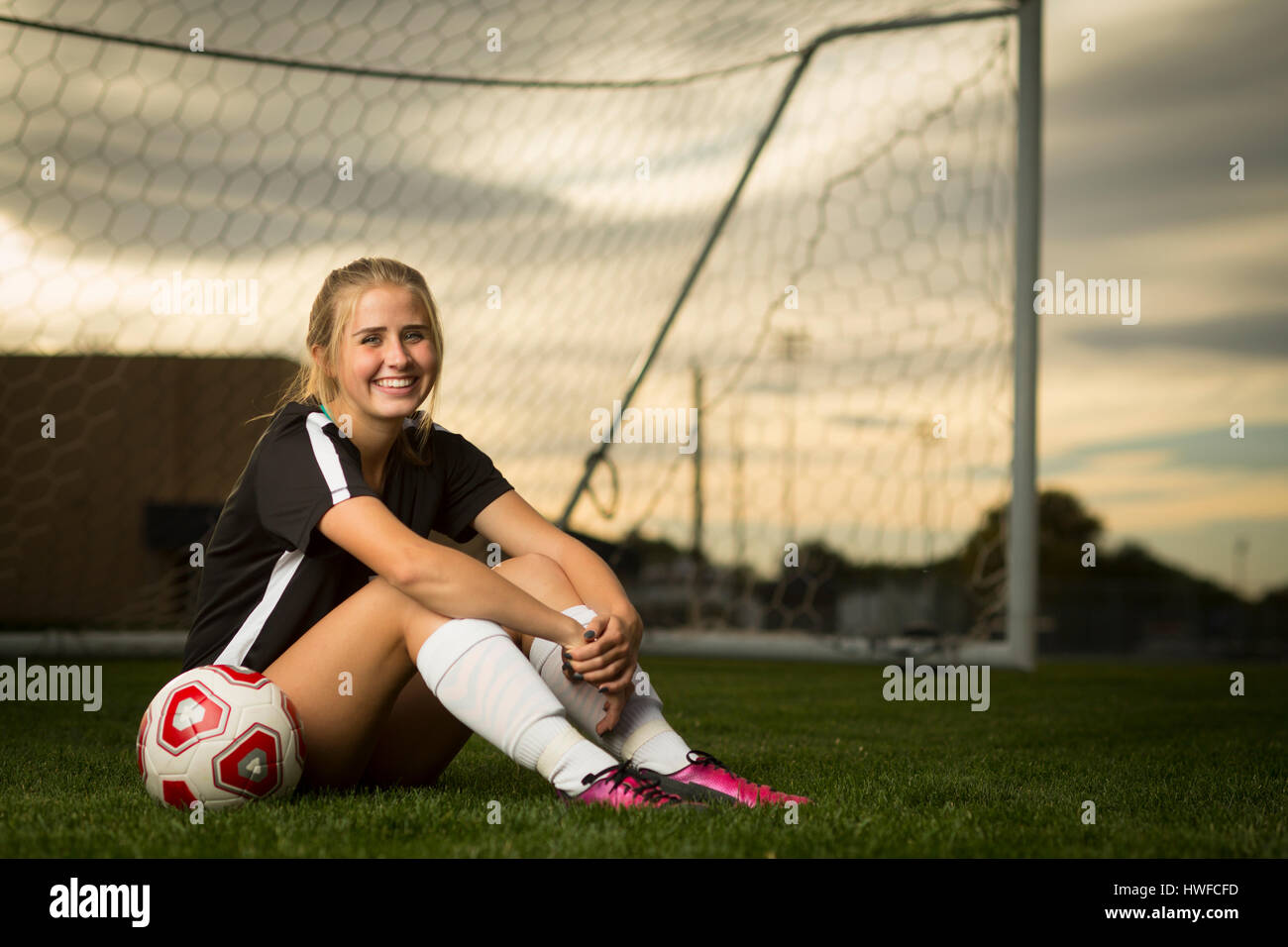 Athlète souriant assis avec balle en buts de soccer sous Ciel de coucher du soleil Banque D'Images